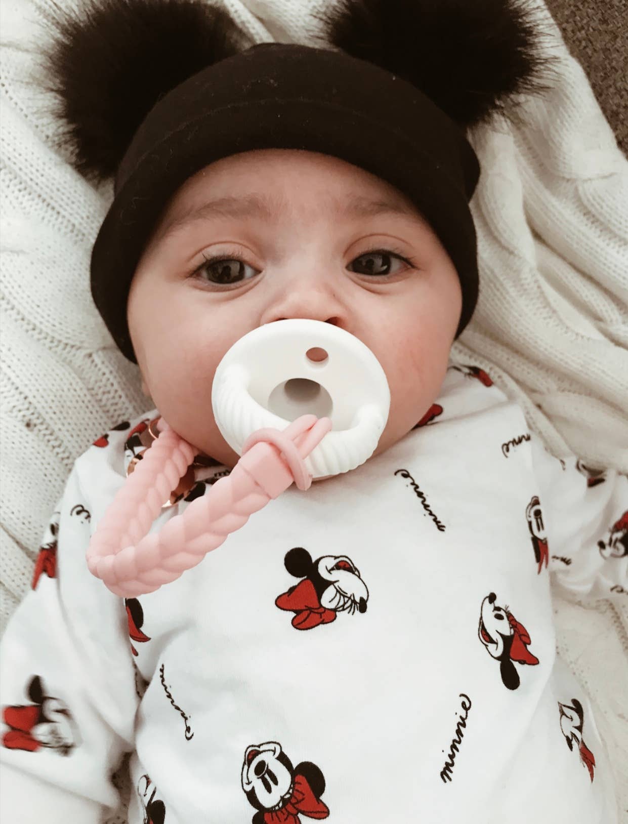 Adorable baby wearing a black beanie and a Mickey Mouse onesie, holding a pacifier with a Sweetie Strap™ clip.
