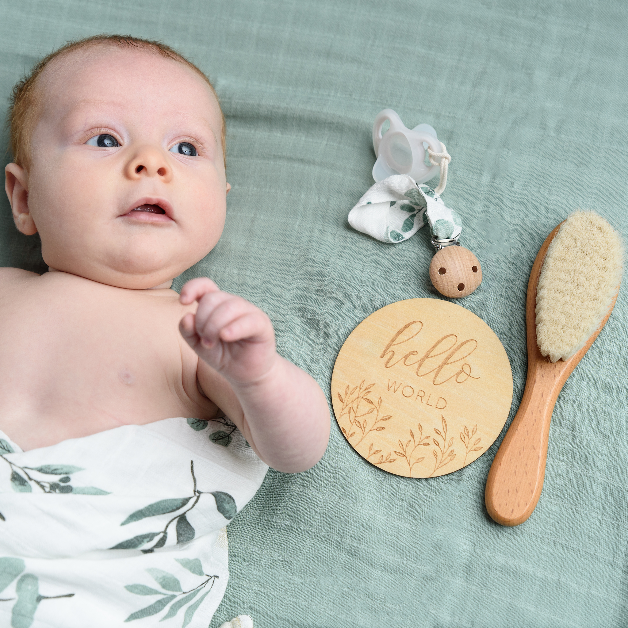 Baby lies on a eucalyptus-themed blanket next to a wooden brush, a "hello world" sign, and a clip, part of a neutral gift set.