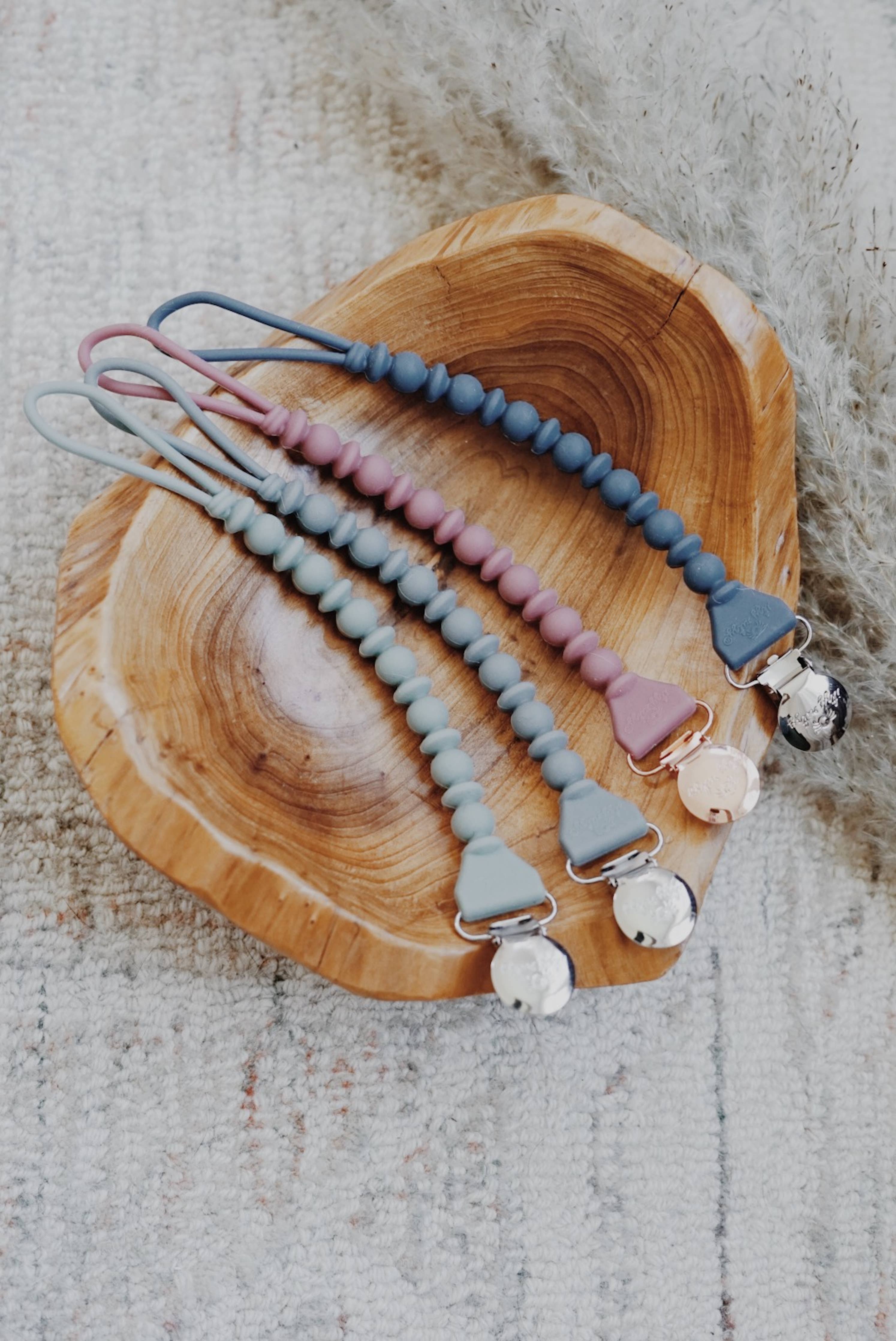 Colorful Sweetie Strap™ Silicone Pacifier Clips displayed on a wooden tray, showcasing braided and beaded designs.