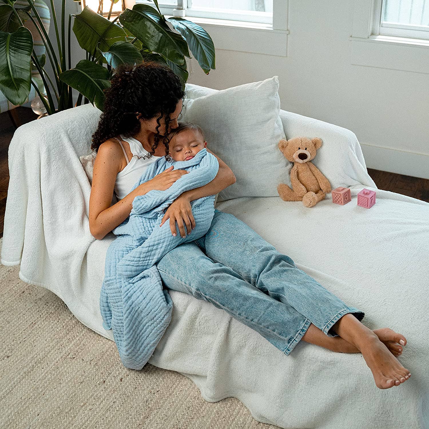 Mom cuddling baby in a blue muslin cotton blanket on a cozy couch, with toys nearby.