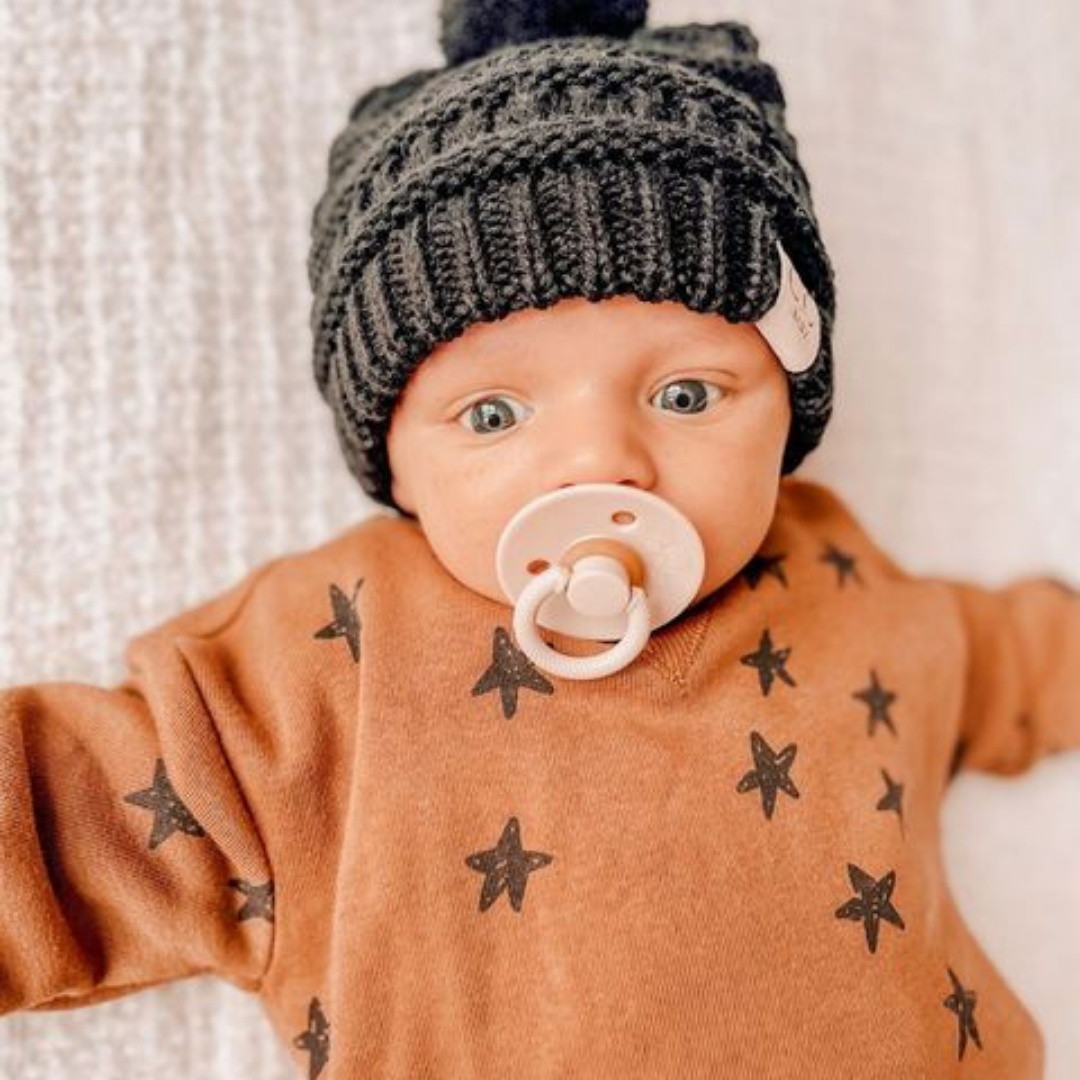 Adorable baby wearing a black knit hat and brown star-patterned shirt, soothing with Itzy Soother natural rubber pacifier.