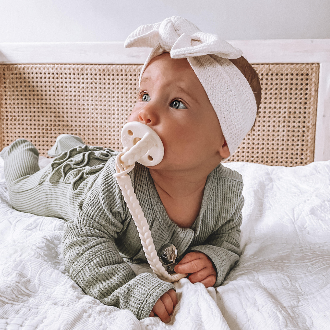 Adorable baby wearing a headband and using a Sweetie Soother™ pacifier, lounging on a cozy bed.