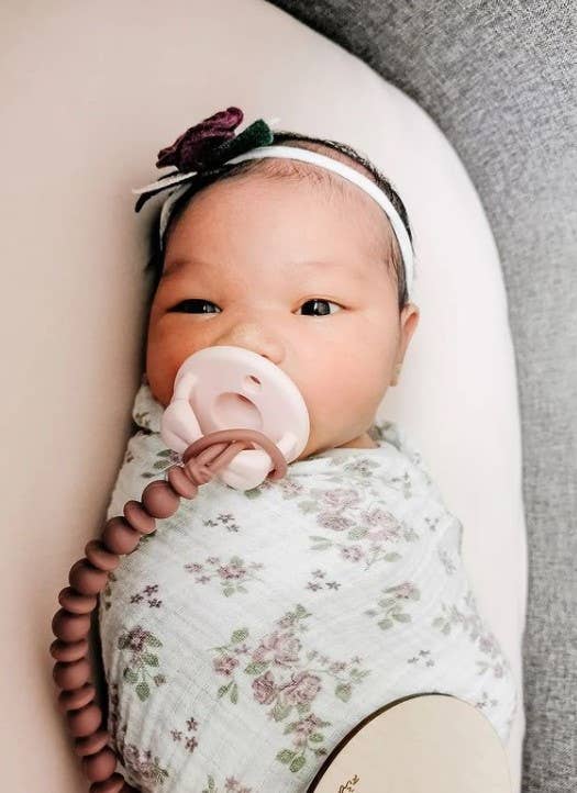 Adorable baby wrapped in floral blanket with Sweetie Strap™ silicone pacifier clip, featuring a pink pacifier.