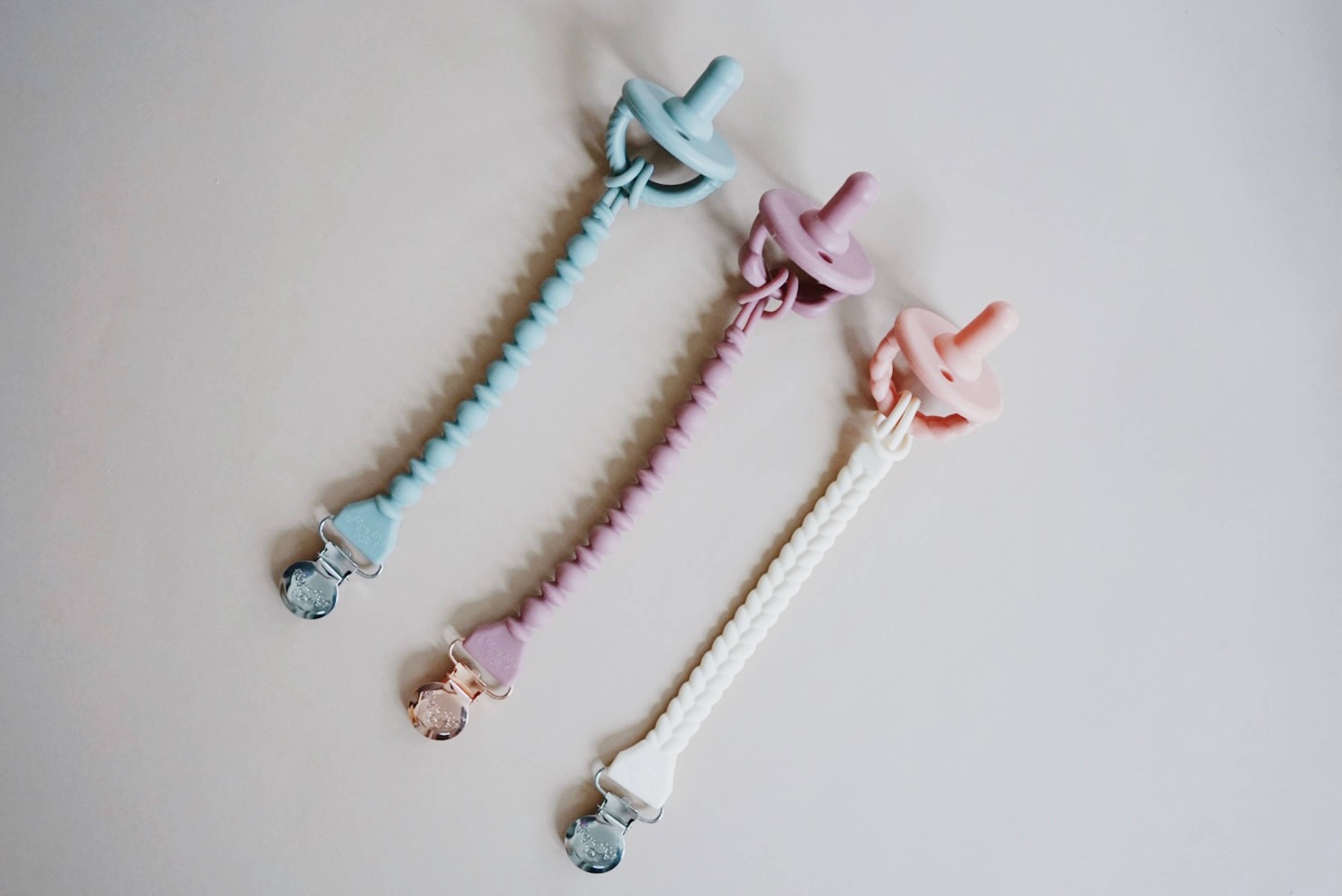 Stylish Sweetie Strap™ silicone pacifier clips in pastel blue, purple, and pink on a neutral background.