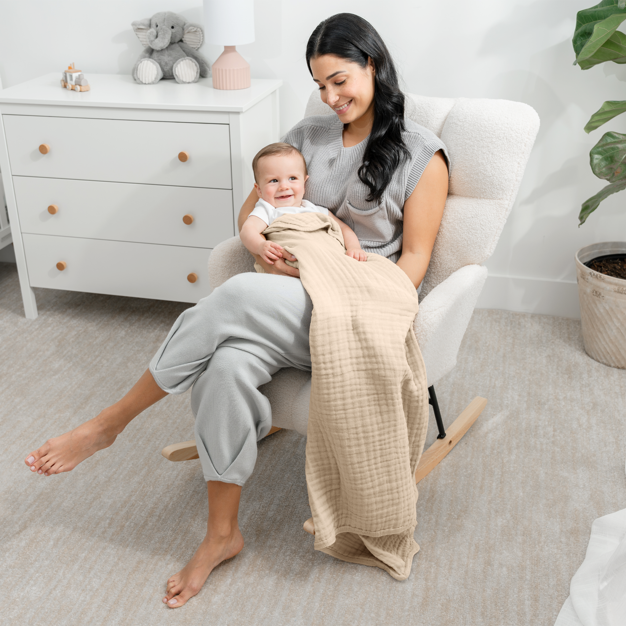 A mother sits in a rocking chair, holding her baby wrapped in a soft muslin cotton blanket by Comfy Cubs.
