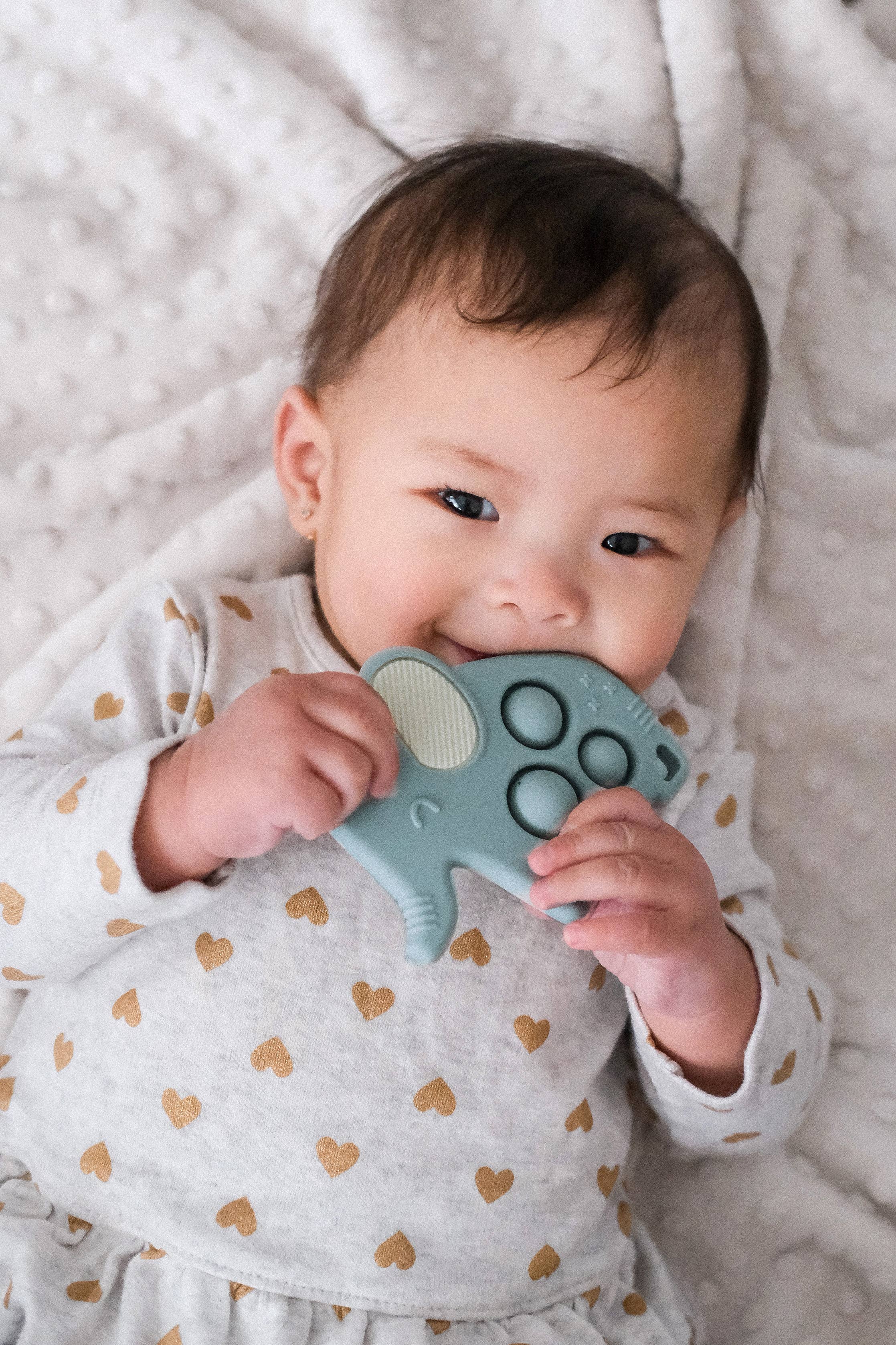 Baby chewing on Itzy Ritzy Itzy Pop Elephant sensory popper toy, wearing heart-patterned outfit on a soft blanket.