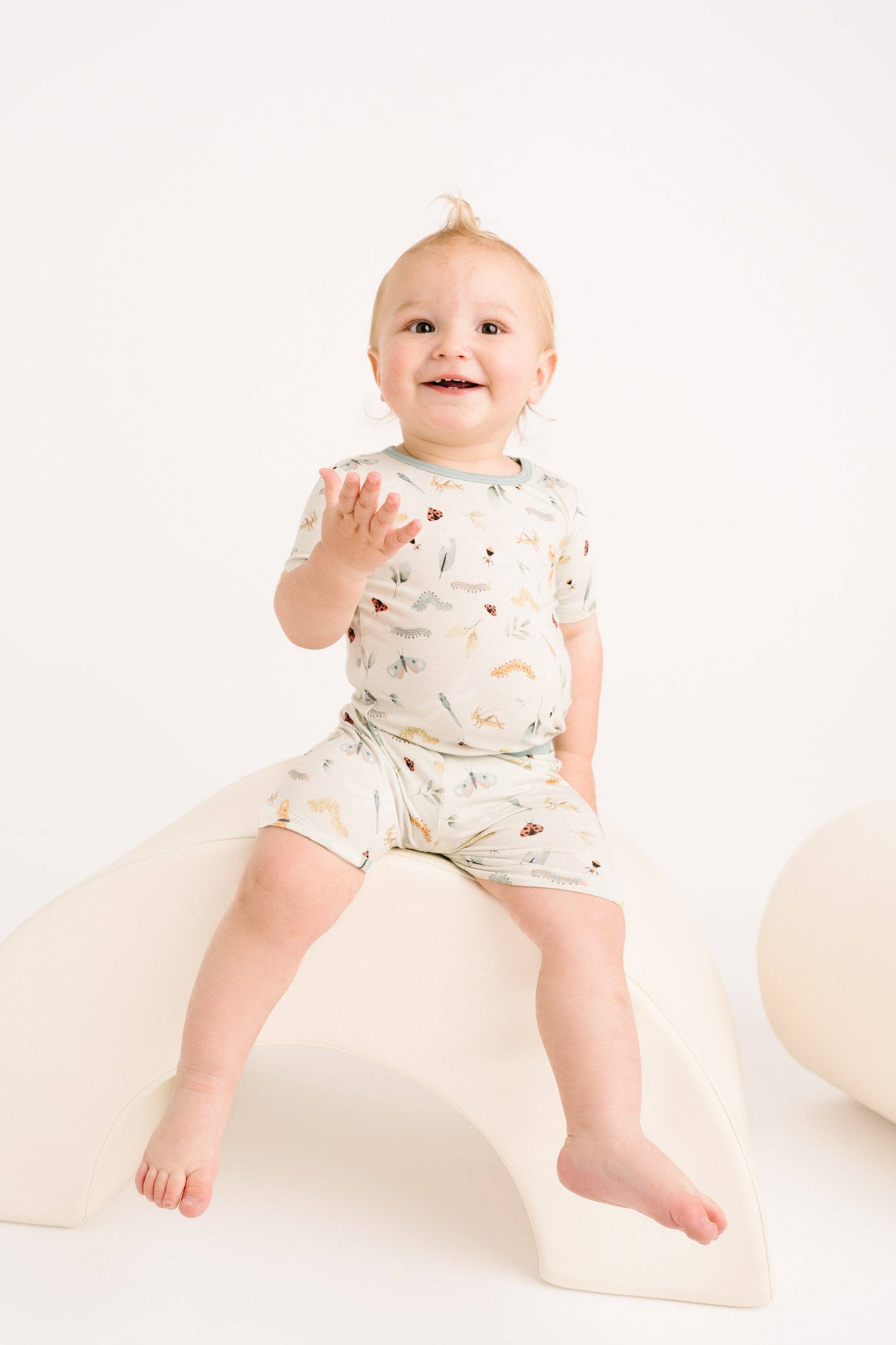 Happy baby wearing Little One Shop Bug Buddies Bamboo Shorts Set, 12-18 months, sitting on a white shape, showcasing comfort and style.