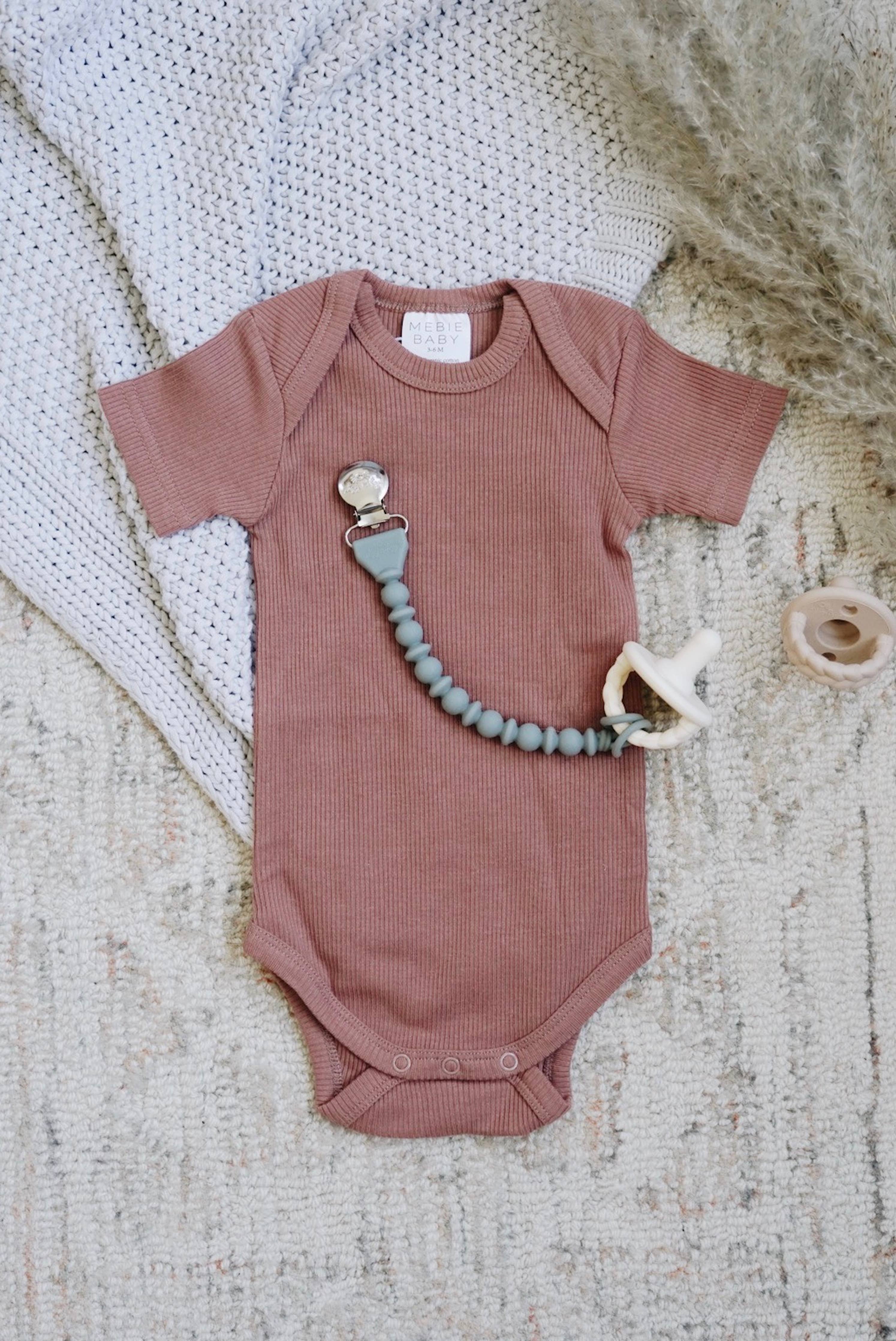 Cute pink baby onesie with a Sweetie Strap™ pacifier clip and wooden teether on a cozy blanket.