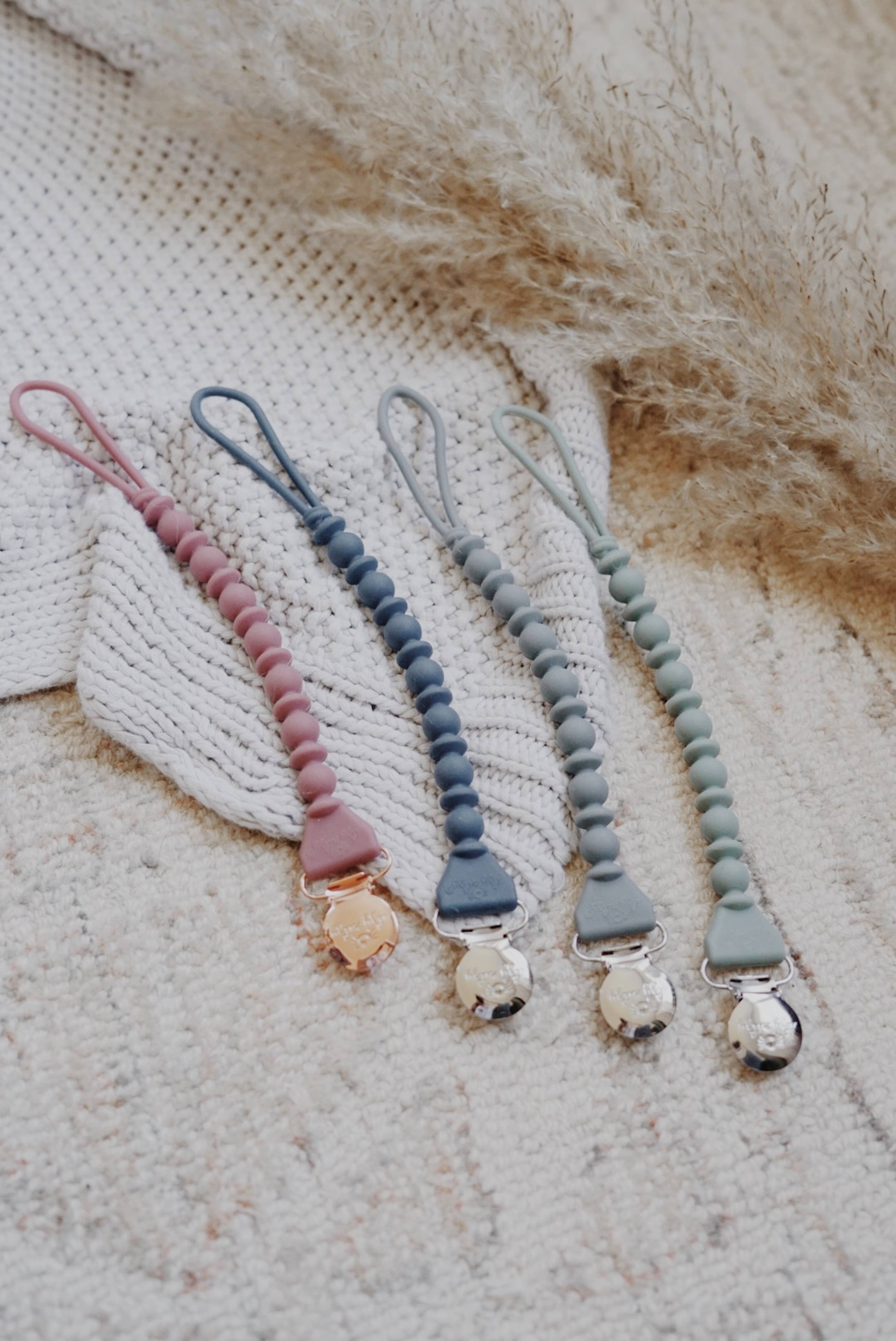 Four stylish Sweetie Strap™ silicone pacifier clips in various colors, showcasing braided and beaded patterns.