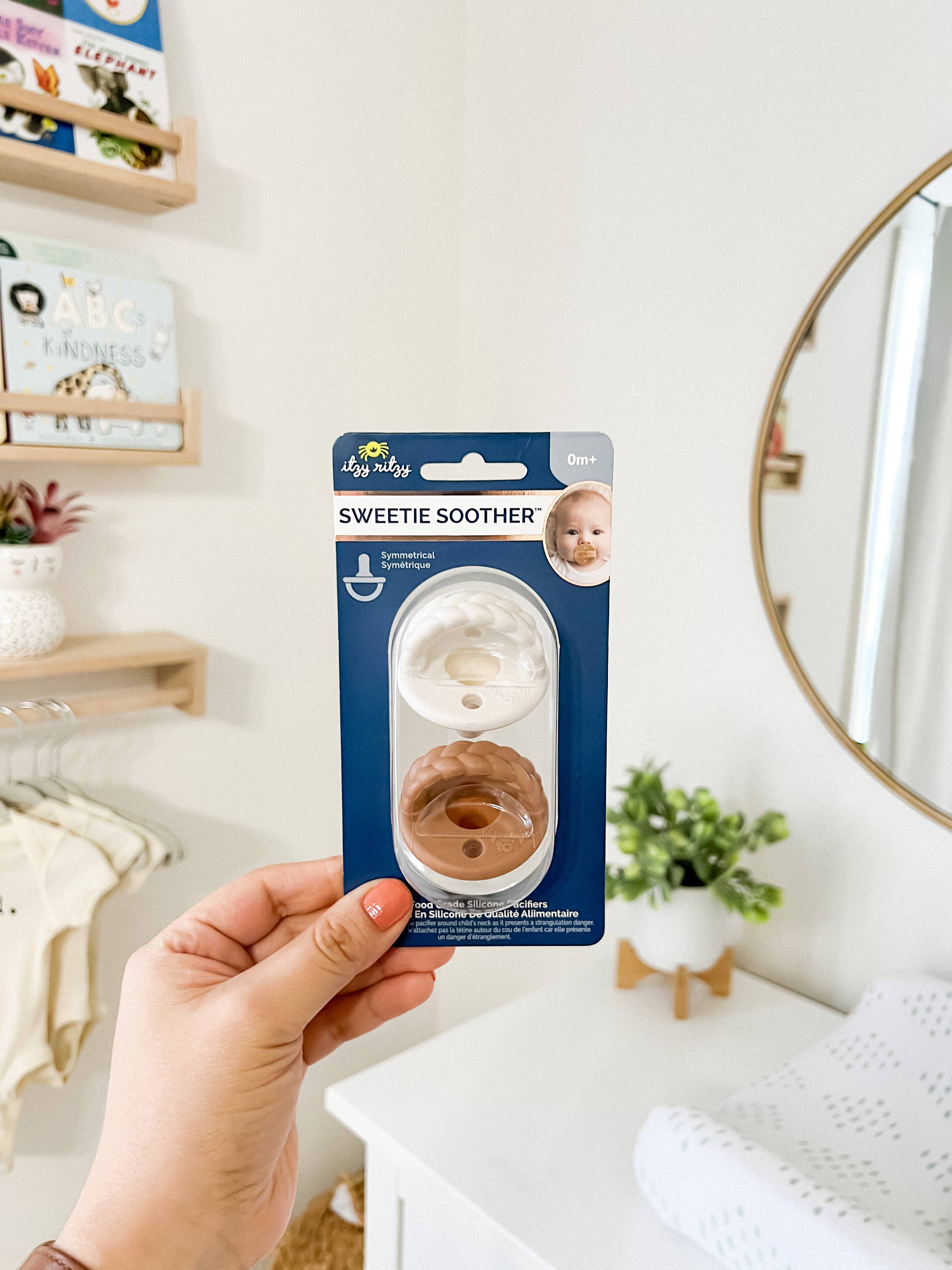 Sweetie Soother™ Pacifier Sets in Toast + Buttercream Braids packaging held in hand against a cozy nursery backdrop.