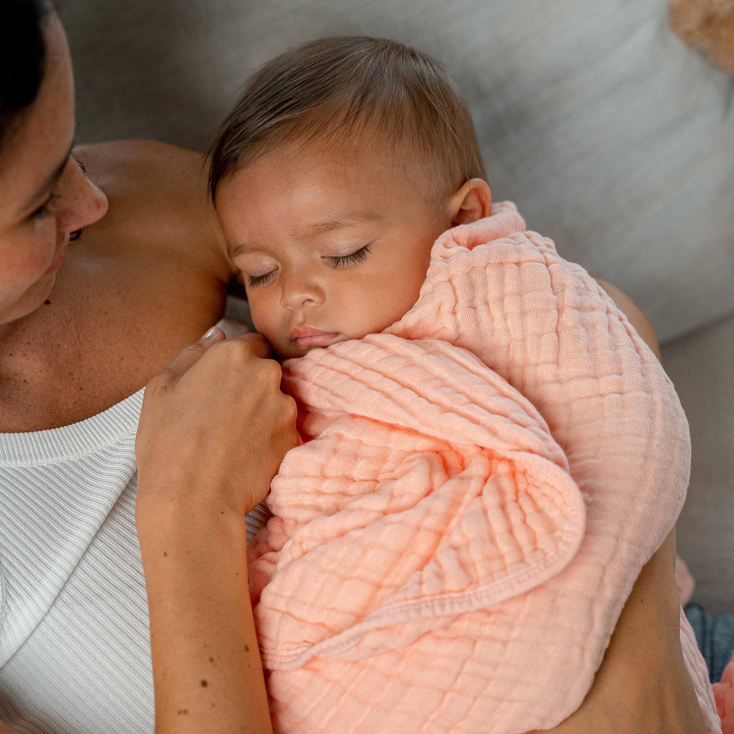 Mom holds a sleeping baby wrapped in a soft peach muslin cotton blanket for ultimate comfort.
