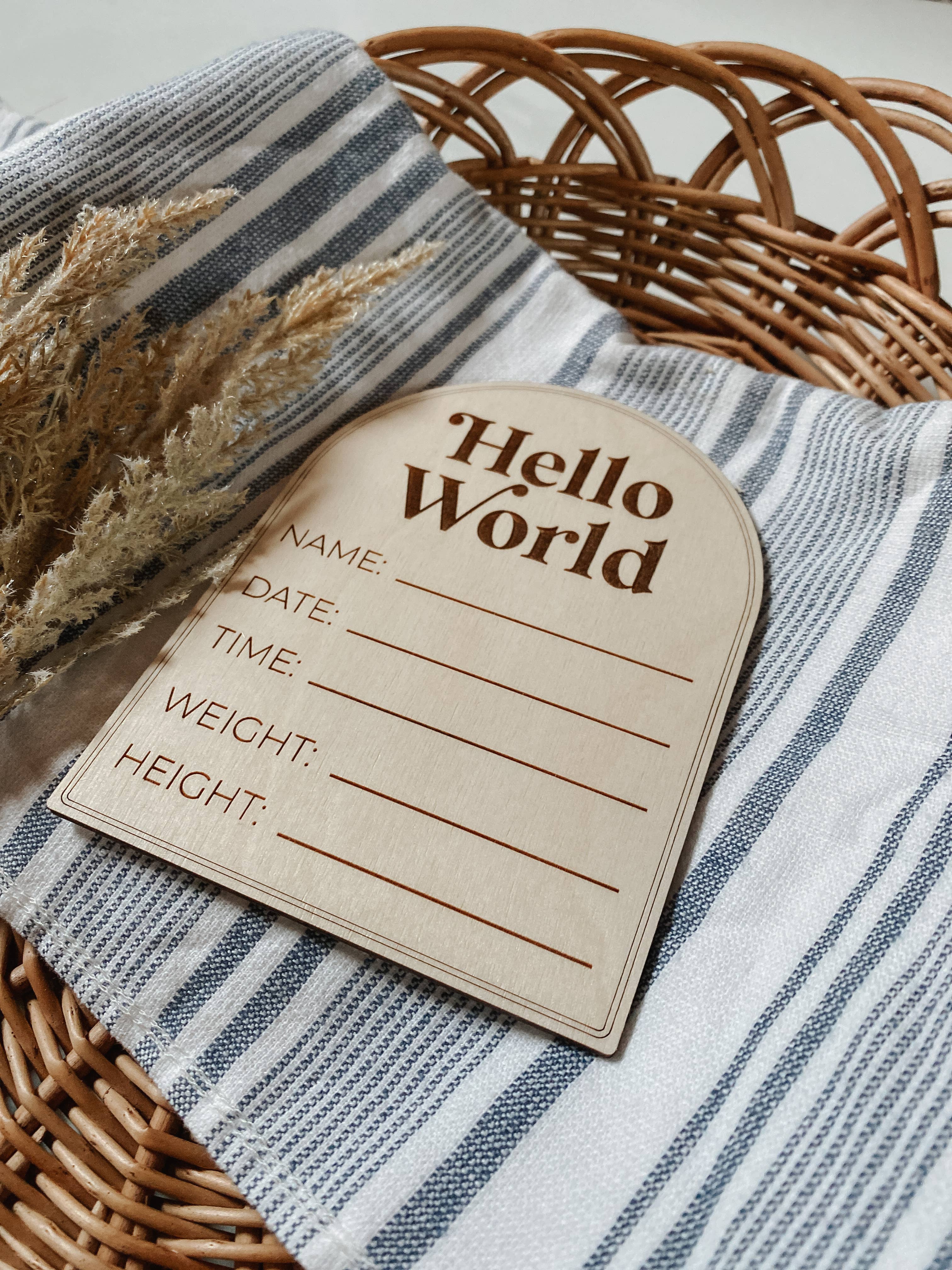 Modern wooden birth announcement plaque with 'Hello World' design, blank spaces for baby's details, placed on fabric.
