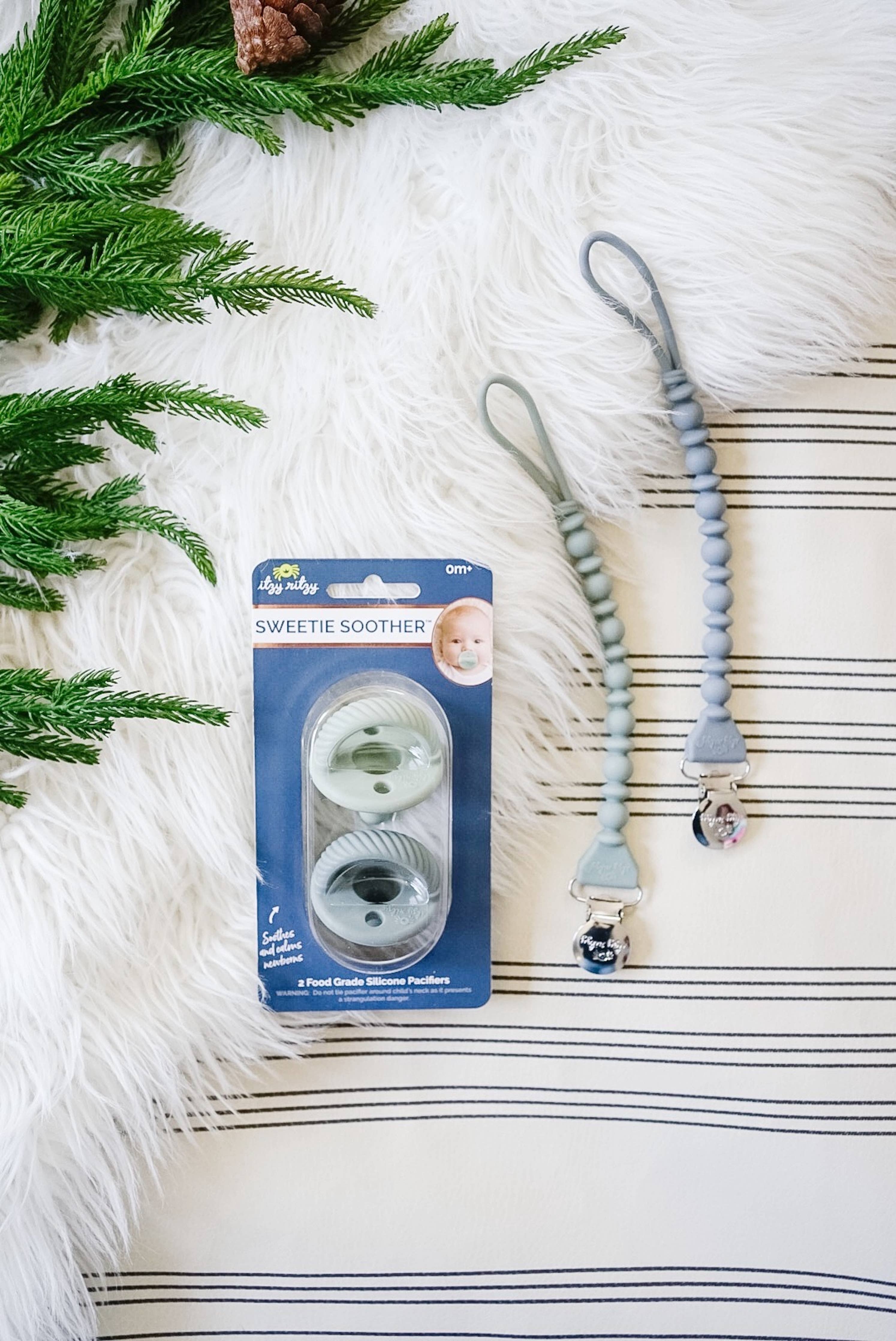 Sweetie Strap™ silicone pacifier clips and Sweetie Soother packaging on a soft, textured background with greenery.